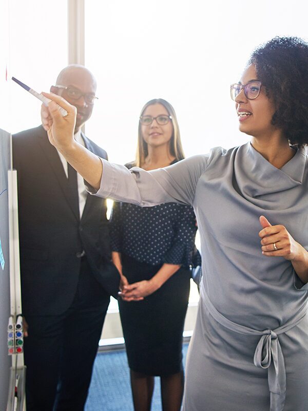 professional woman pointing at whiteboard while others look.