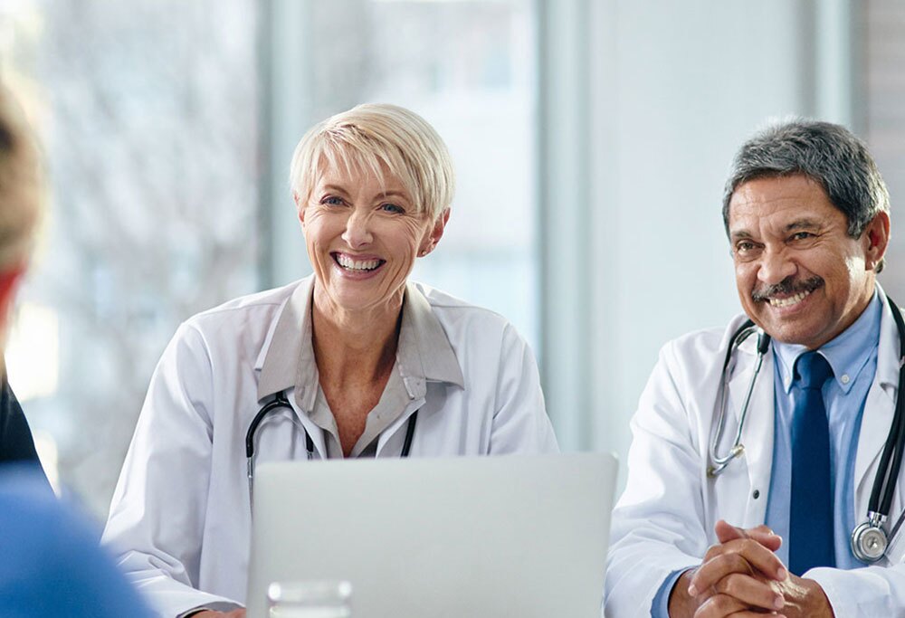 Two doctors seated at a table talking and smiling with somebody off camera.